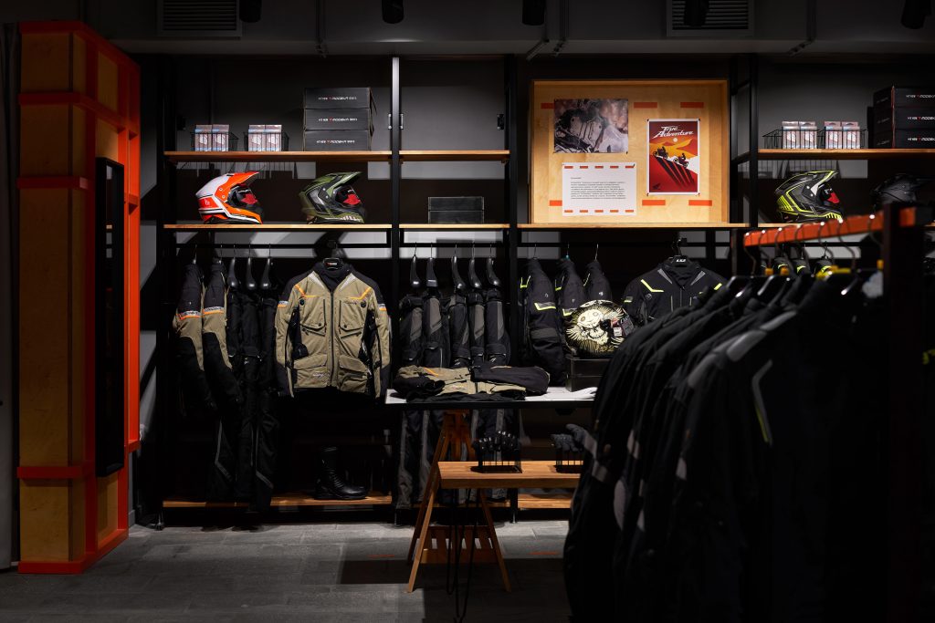 Мотосалон с оранжевыми акцентами, графикой и мотоэкипировкой на торговом оборудовании.