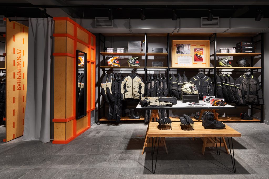 Торговый зал мотосалона и примерочная с экипировкой на серой стене и напольное торговое оборудование из дерева на заказ.