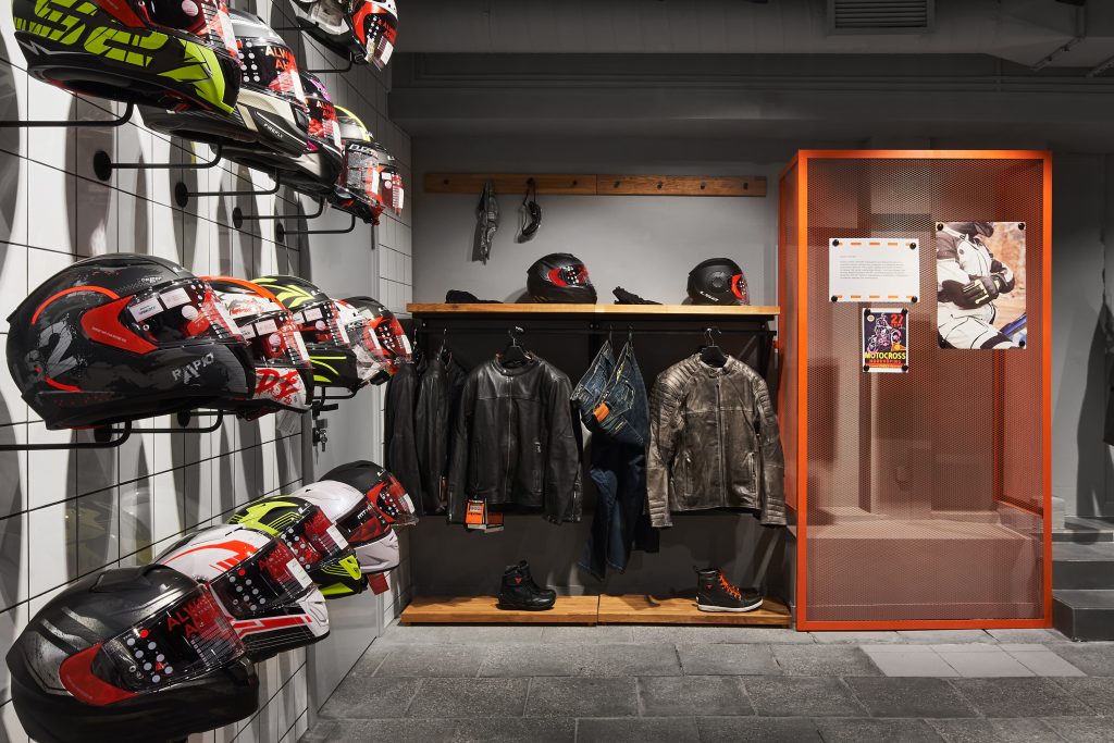 Мотосалон с оранжевыми постерами мотоциклов на рифленой металлической стене и дизайнерское торговое оборудование.