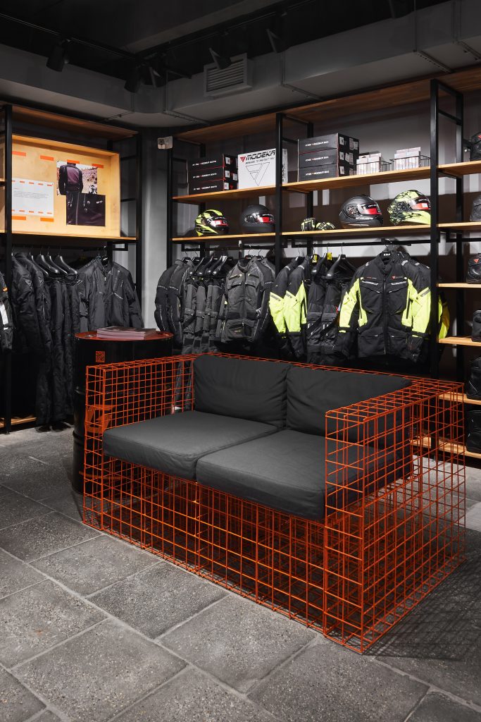 Зона одыха в мотосалоне. Диван из оранжевой металлической сетки и мягкие подушки. Торговое оборудование и мотоэкипировка.